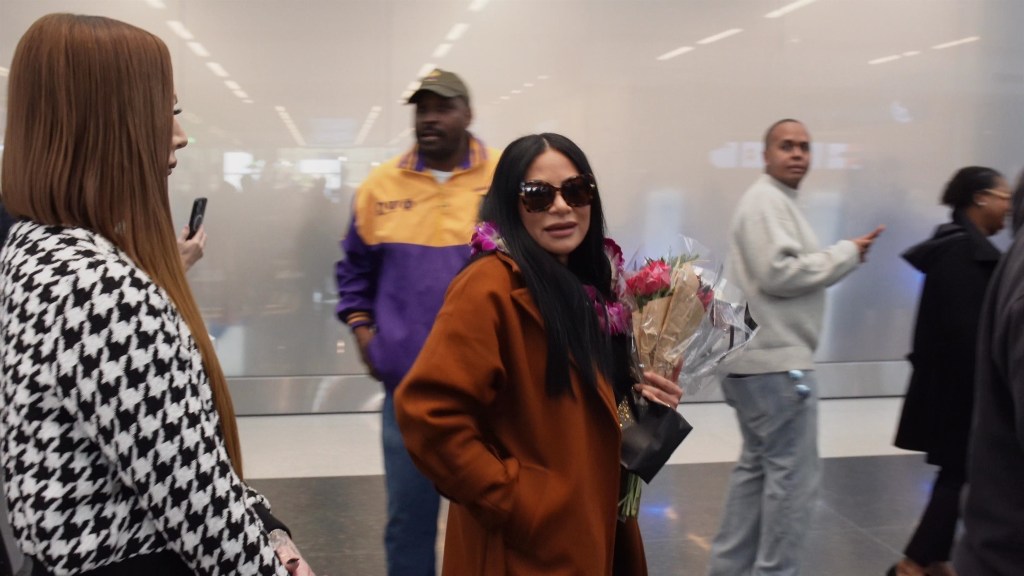 Jen Shah wearing sunglasses, a brown coat, and a lei, holding a bouquet of flowers, at Salt Lake City International Airport.