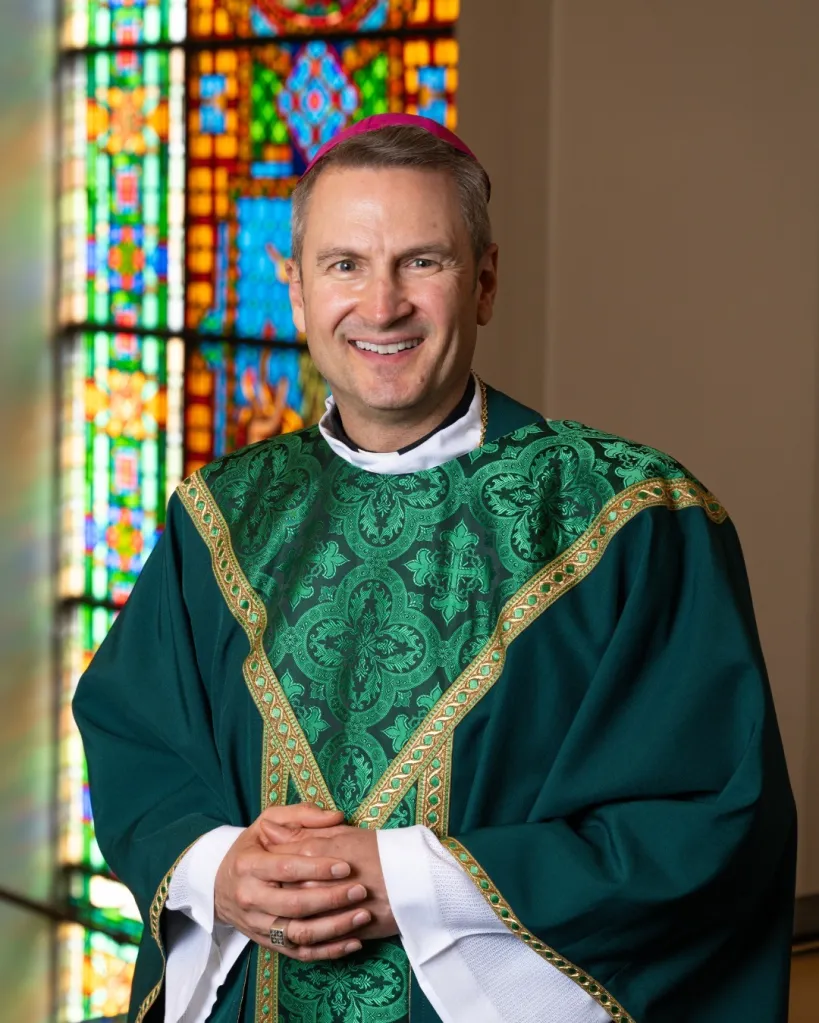 Bishop Ronald Hicks smiling in a green chasuble with a stained glass window behind him.