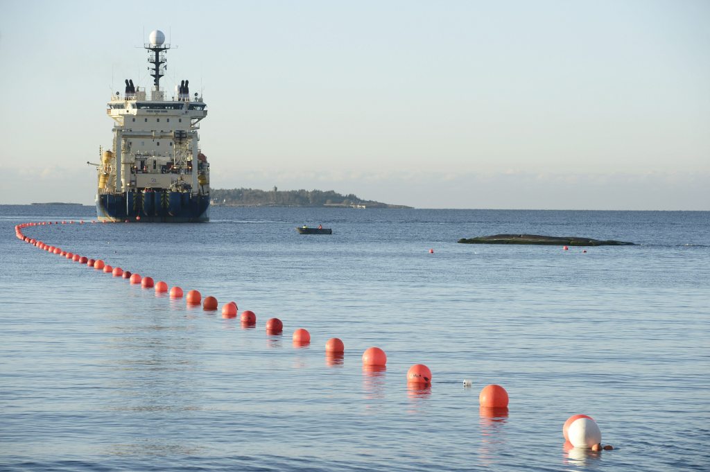A large cable-laying ship in the Baltic Sea, with a line of orange buoys stretching from the ship towards the foreground, indicating the path of a newly laid submarine telecommunications cable.