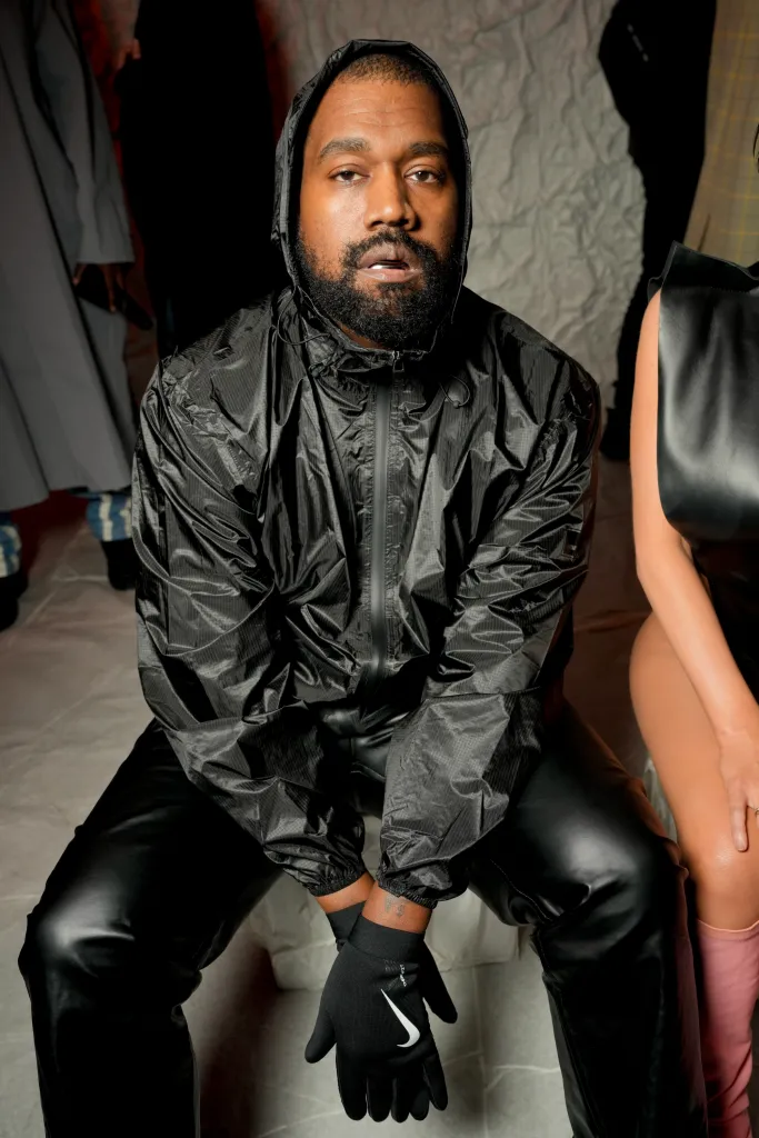 Kanye West sitting in a black hooded jacket, black leather pants, and black Nike gloves, looking directly at the camera.