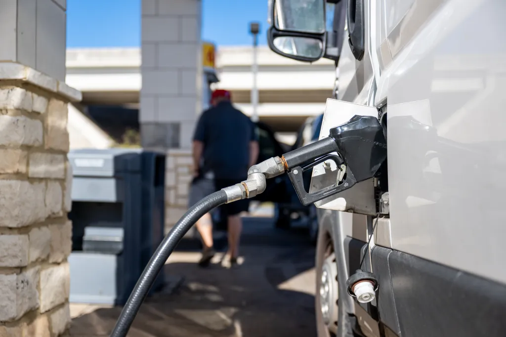 A gas pump nozzle in a vehicle, with a man blurred in the background.