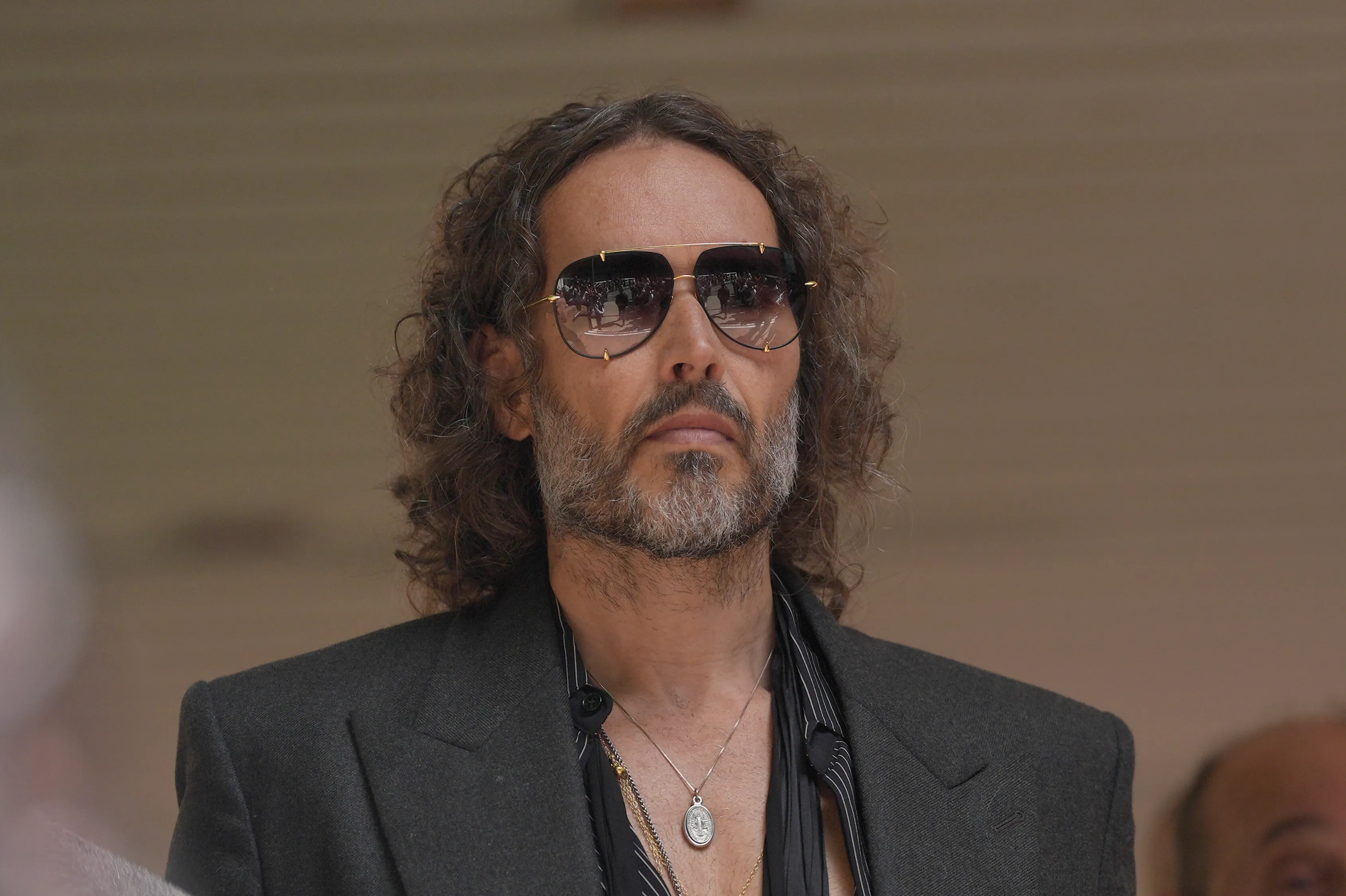 Russell Brand looking forward, wearing sunglasses that reflect a crowd.