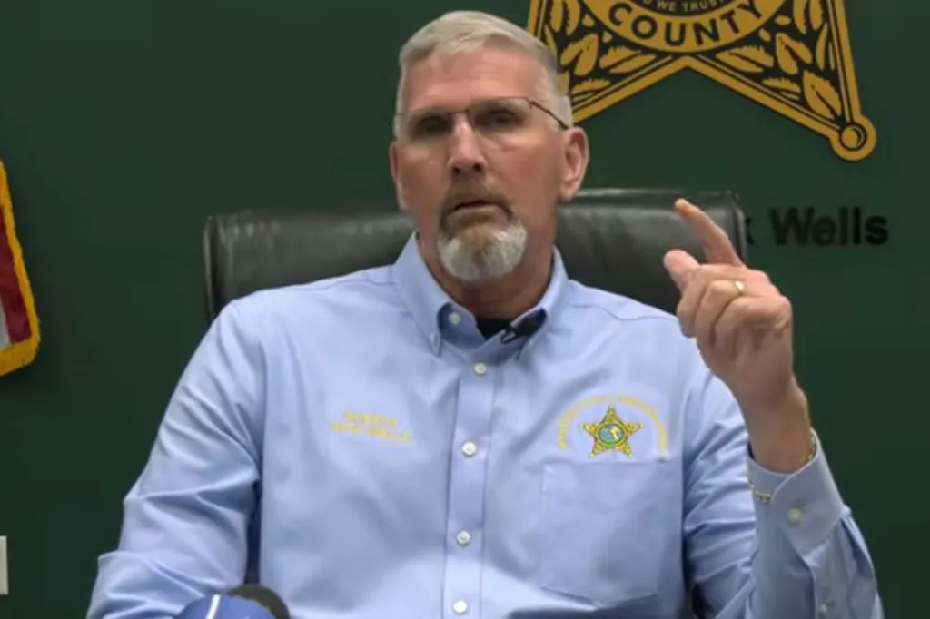 Man with gray hair and beard wearing a light blue shirt with a sheriff's badge speaking at a news conference.