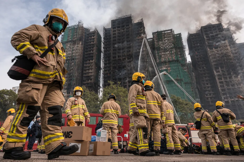 Firefighters in protective gear stand near burnt high-rise buildings, some still smoking, with others spraying water.