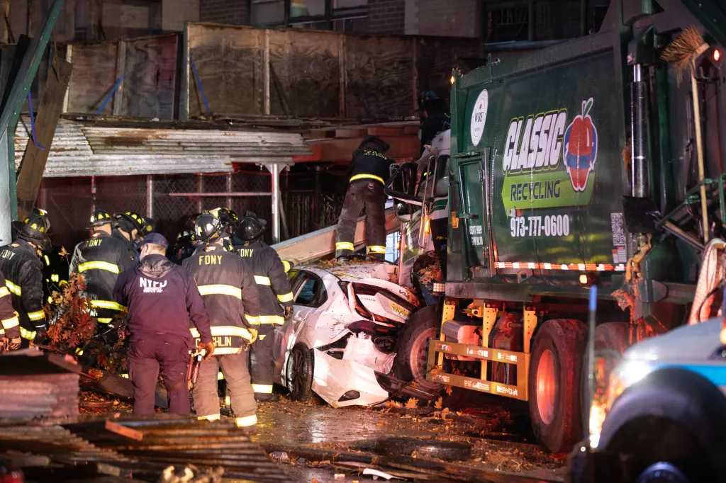 Firefighters and police at the scene of a crash involving a private garbage truck, a white car, and scaffolding.