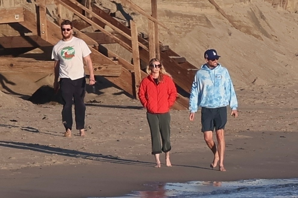 Jake Reiner, Maria Gilfillan, and Alex Silberberg walk barefoot on a beach.