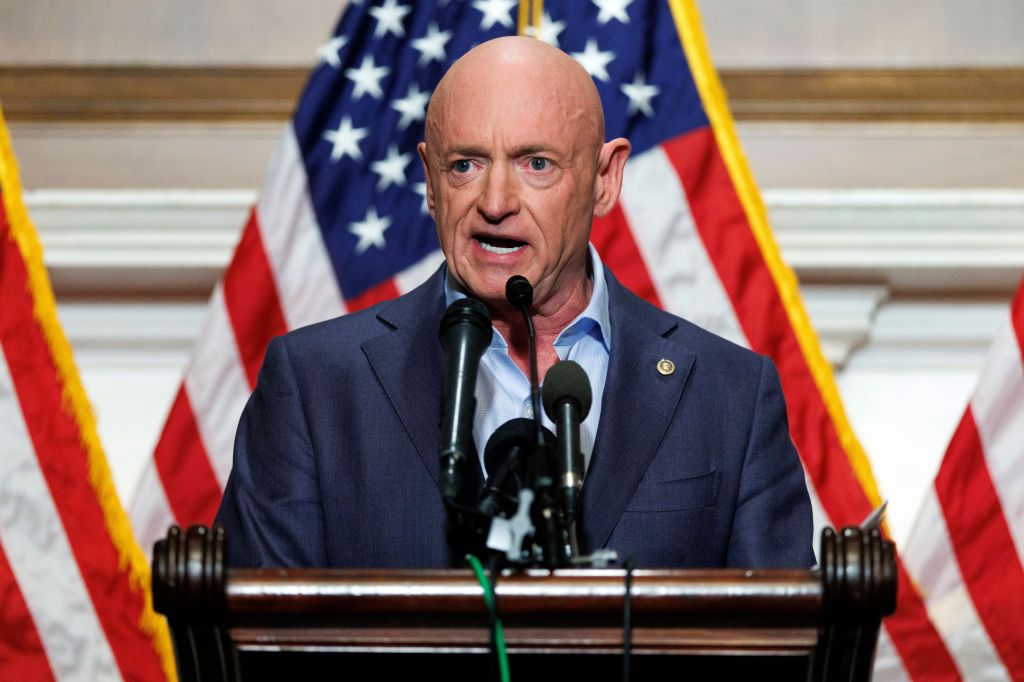 Senator Mark Kelly speaking at a press conference with American flags in the background.