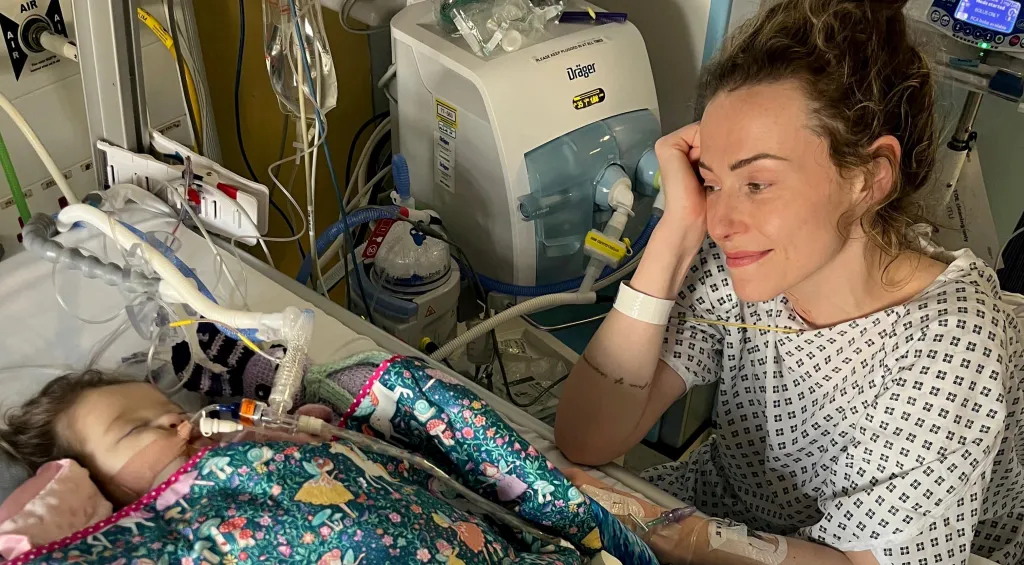 Elle Daniel looks at her daughter Ruby in a hospital, where Ruby is connected to medical equipment.