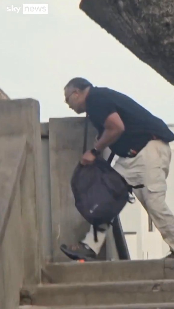 A man in sunglasses, a black shirt, and khaki pants walks down concrete stairs, holding a black backpack.
