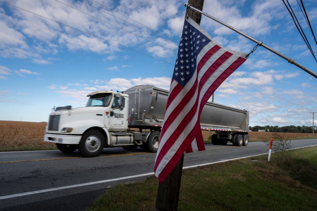US flag on a pole in the foreground with a truck driving down a rural road in the background.