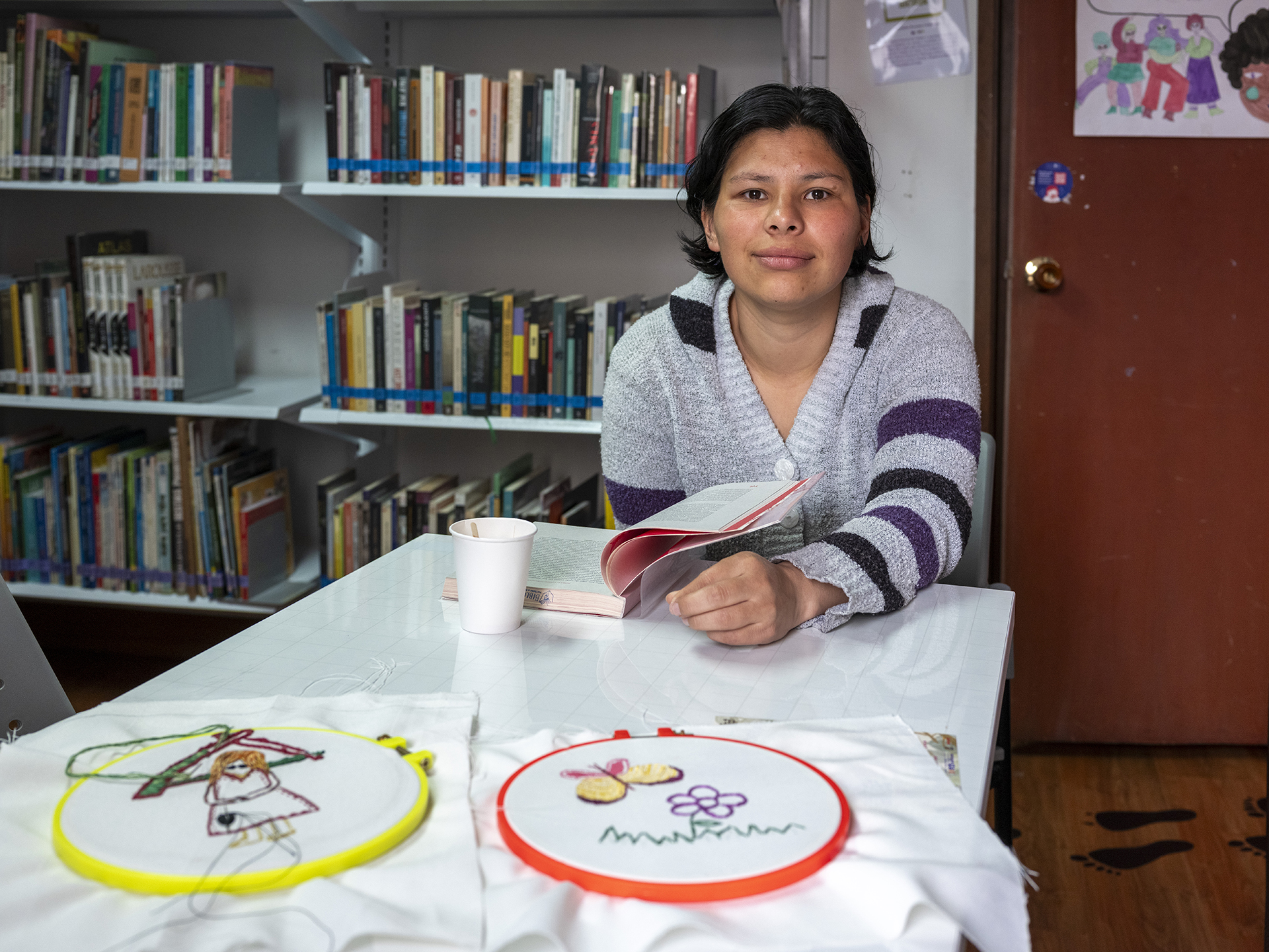 a young woman is reading a book in a classroom in front of children’s in-progress embroidery projects. A large bookshelf is just behind her.