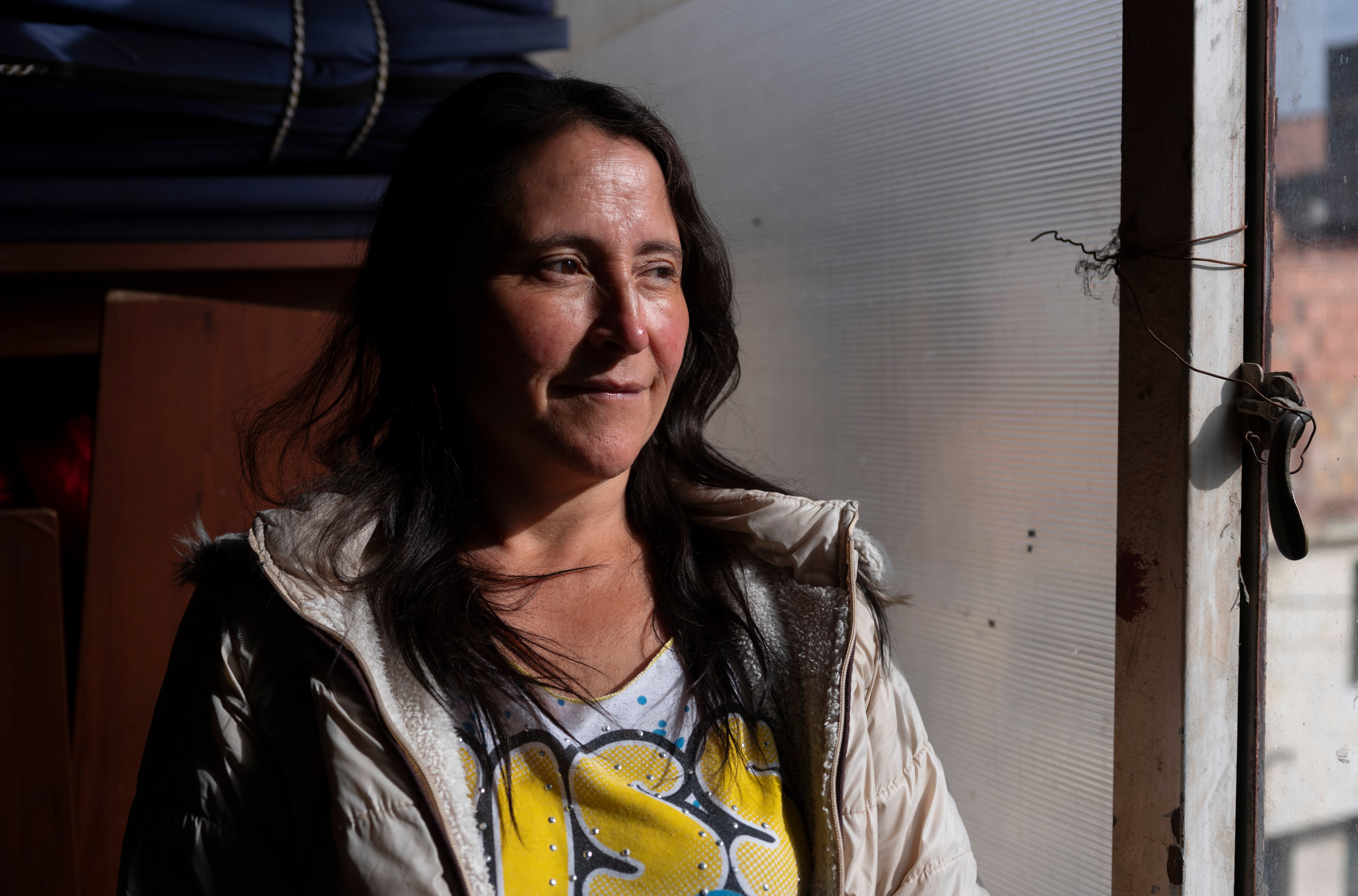 a woman with a dark shadow falling over her face looks out a window with a slight smile.