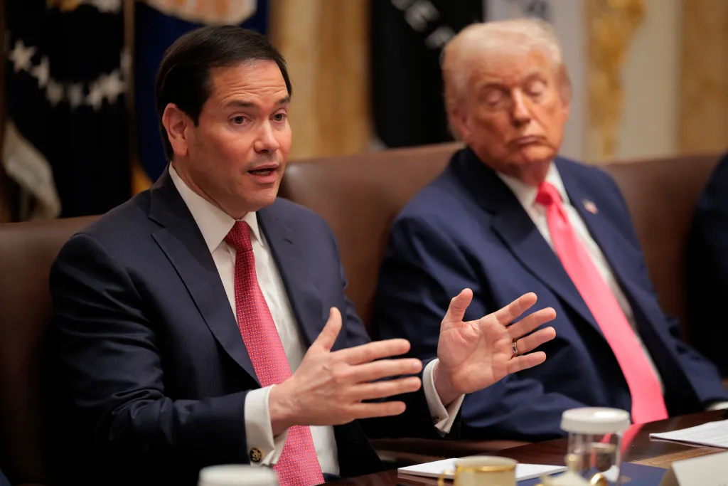 U.S. Secretary of State Marco Rubio speaking with Donald Trump in the background.