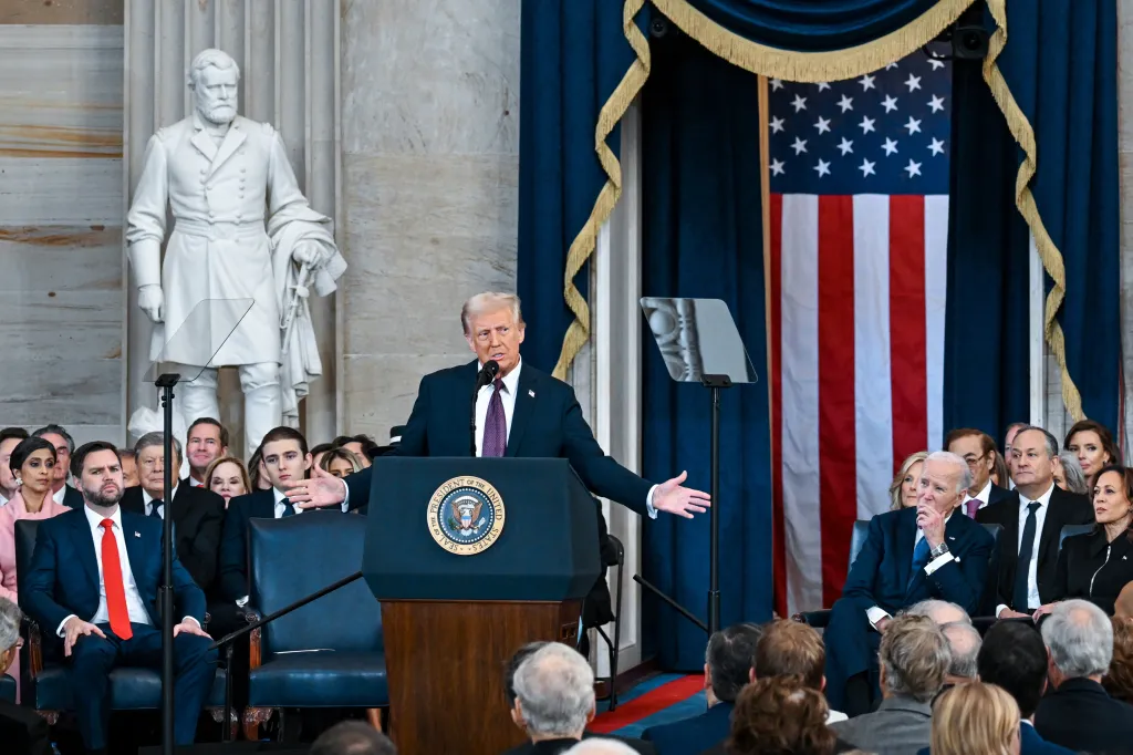 Donald Trump speaking at a podium with the Seal of the President of the United States on it, flanked by a U.S. flag and a statue of Ulysses S. Grant, with Joe Biden and other dignitaries in the audience.