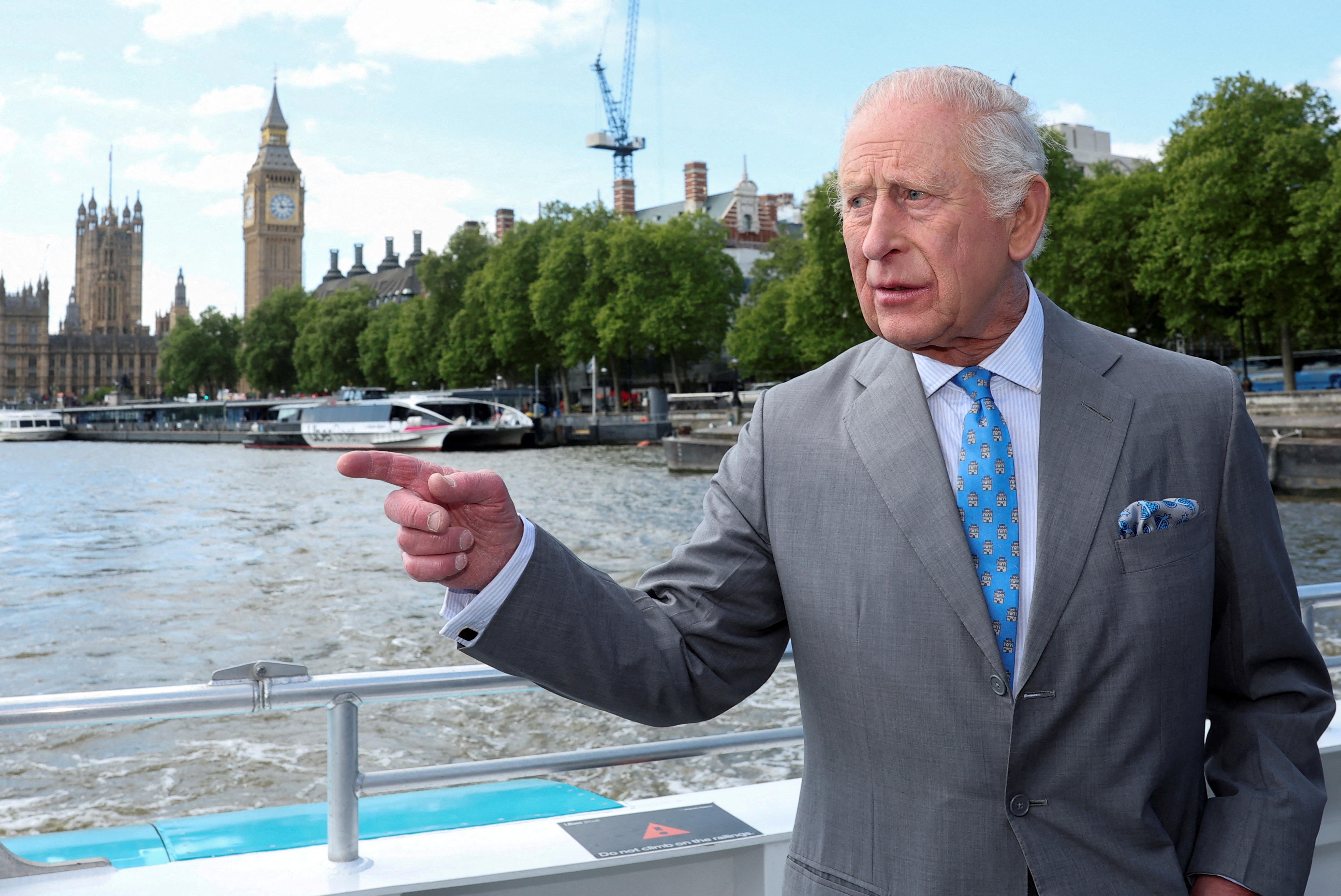 King Charles III gestures from a boat on the River Thames with the Houses of Parliament in the background.