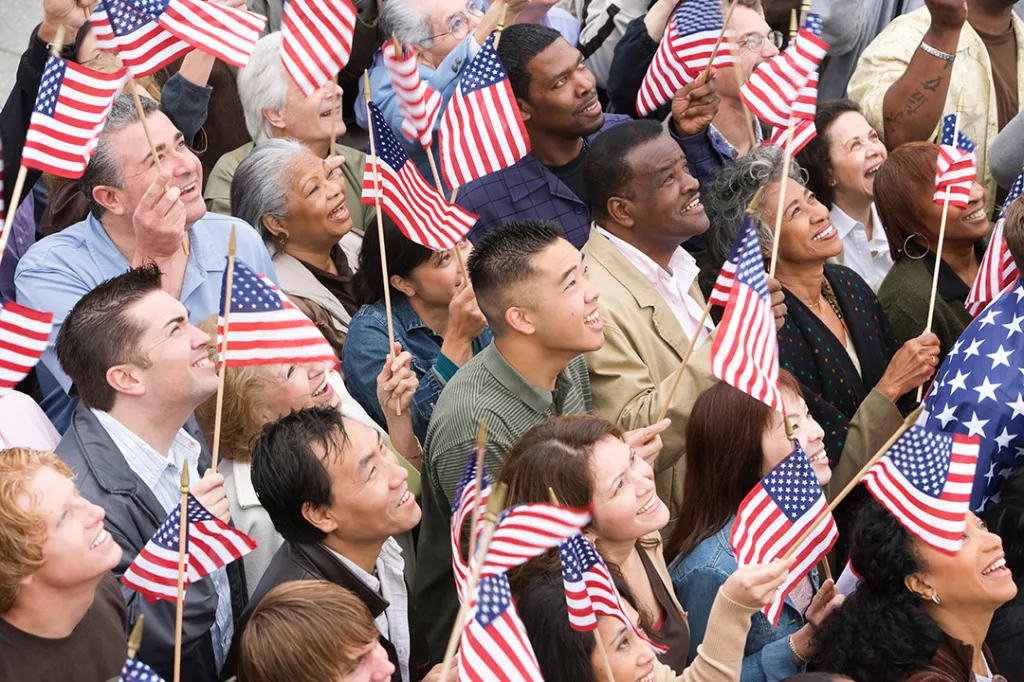 A diverse crowd of people holding small American flags and looking up, smiling.