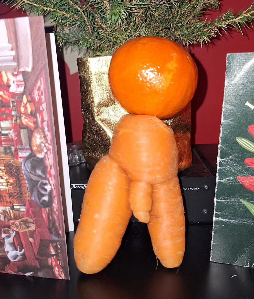 A suggestive carrot resembling a man's anatomy with an orange placed on top, standing near Christmas decorations.