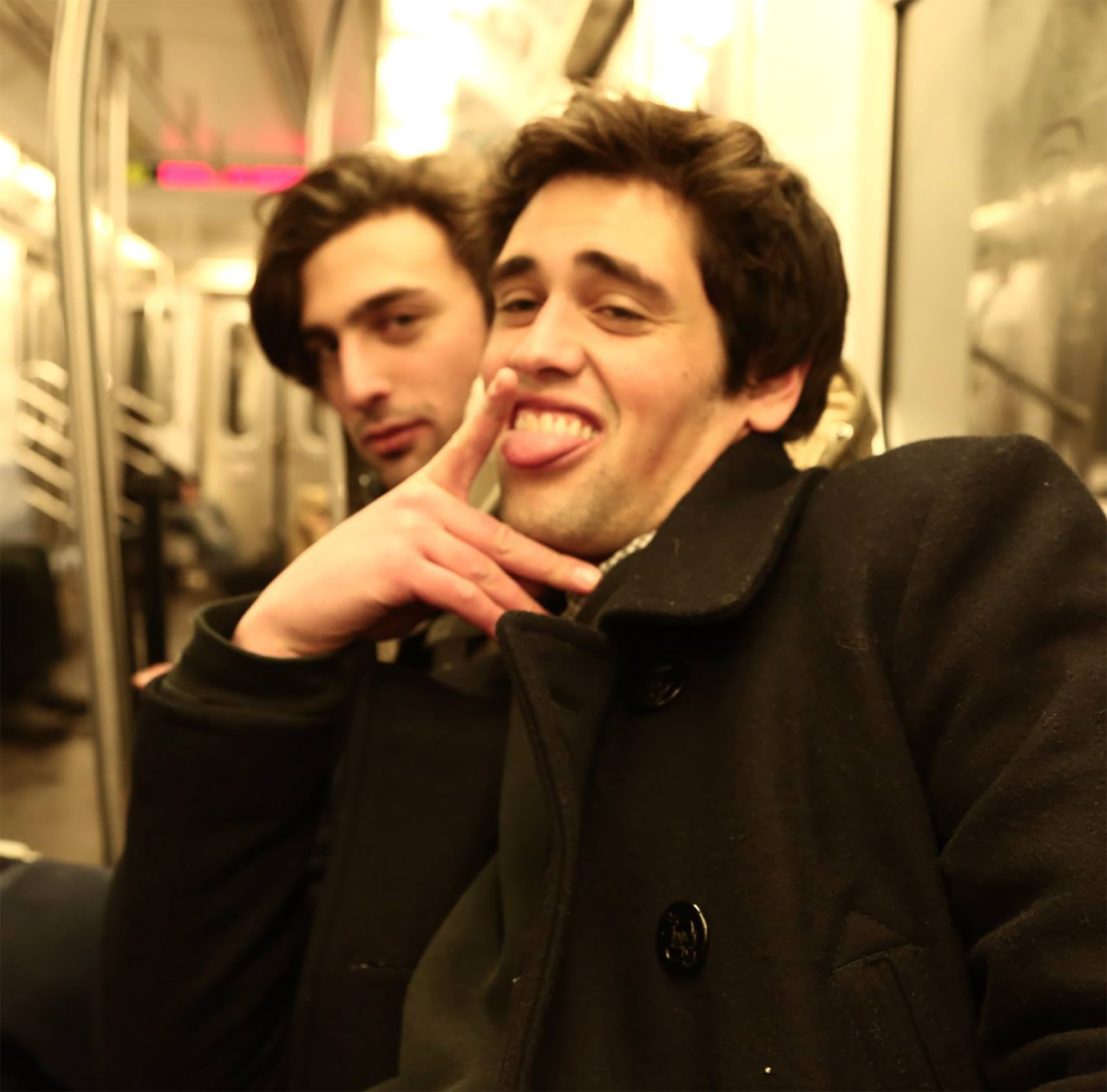 Two men on a train, one making a funny face with his tongue out.