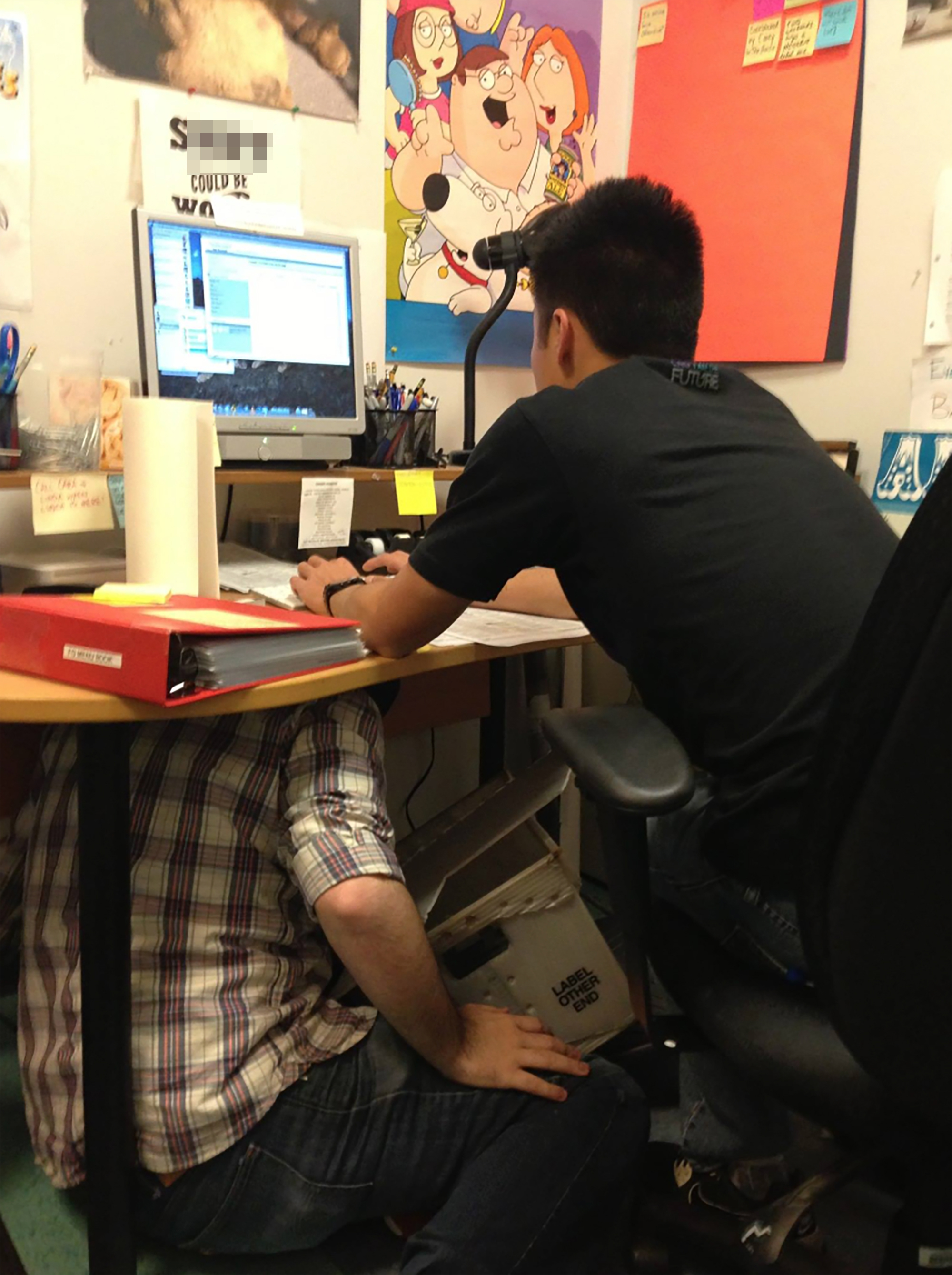 Two men sitting at a desk, one working on a computer and the other crouching underneath the desk.