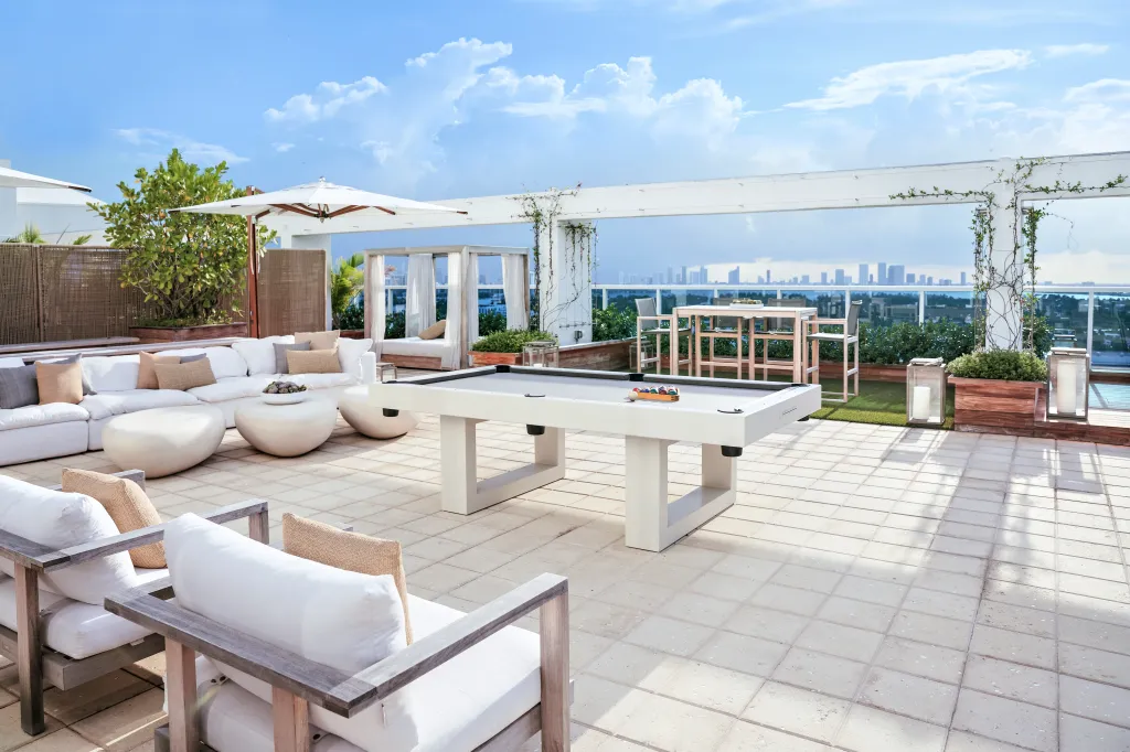 A luxurious rooftop terrace with a pool table, lounge seating, and distant views of the Miami skyline.