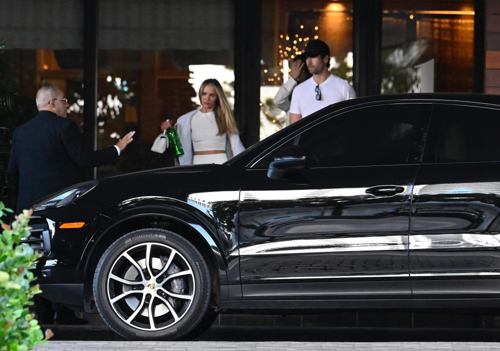 Glen Powell opening the door of a black SUV for Michelle Randolph at a hotel.