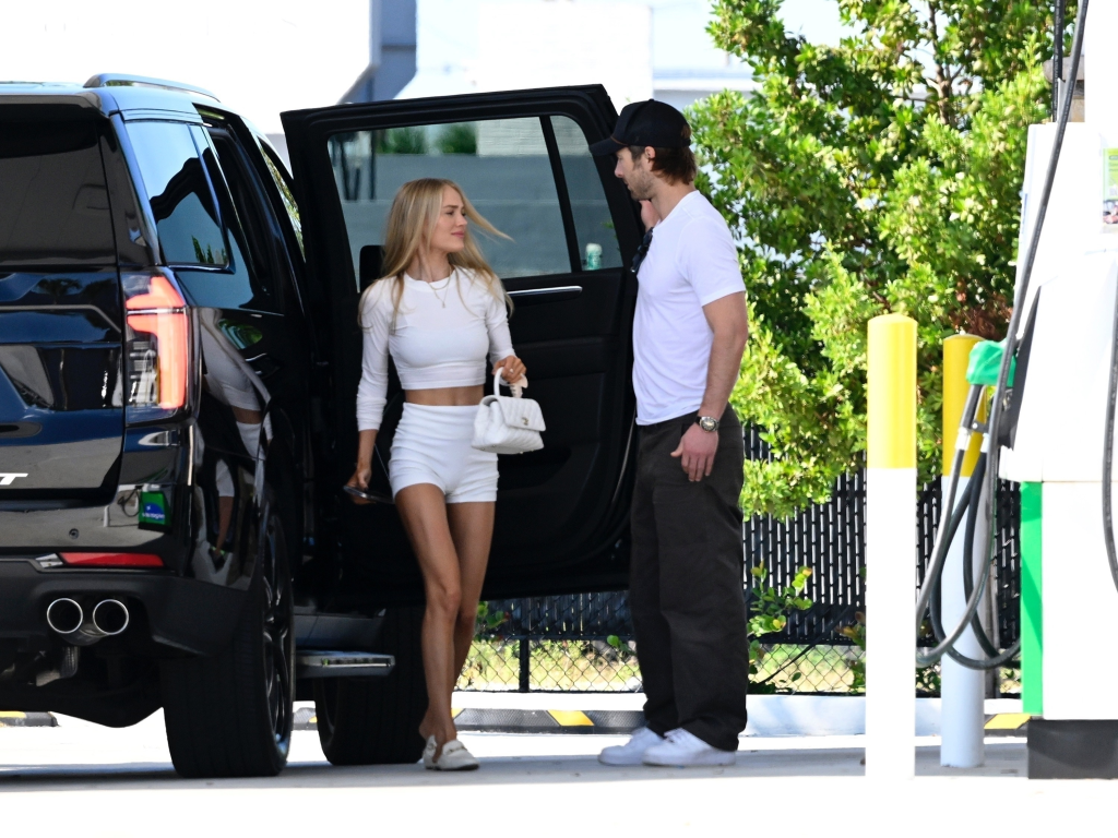 Glen Powell holding a car door open for Michelle Randolph at a gas station.