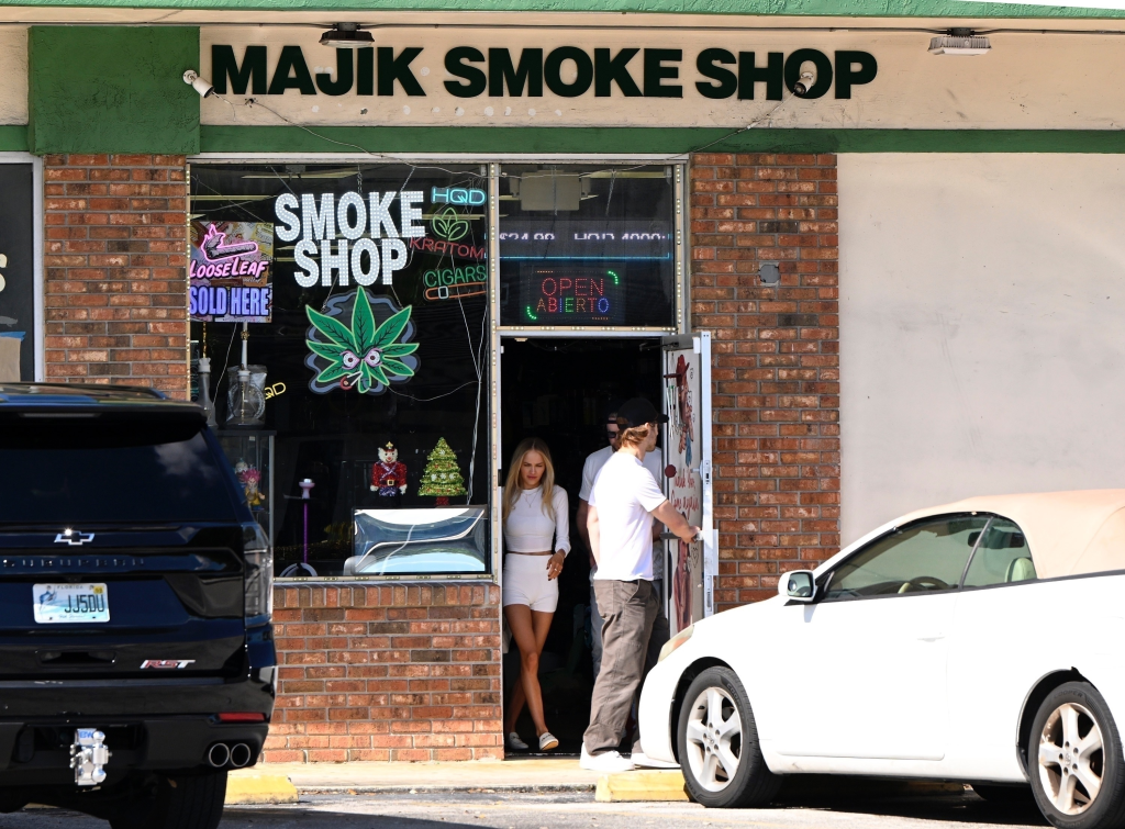 Glen Powell holding a door open for Michelle Randolph as they leave a smoke shop.