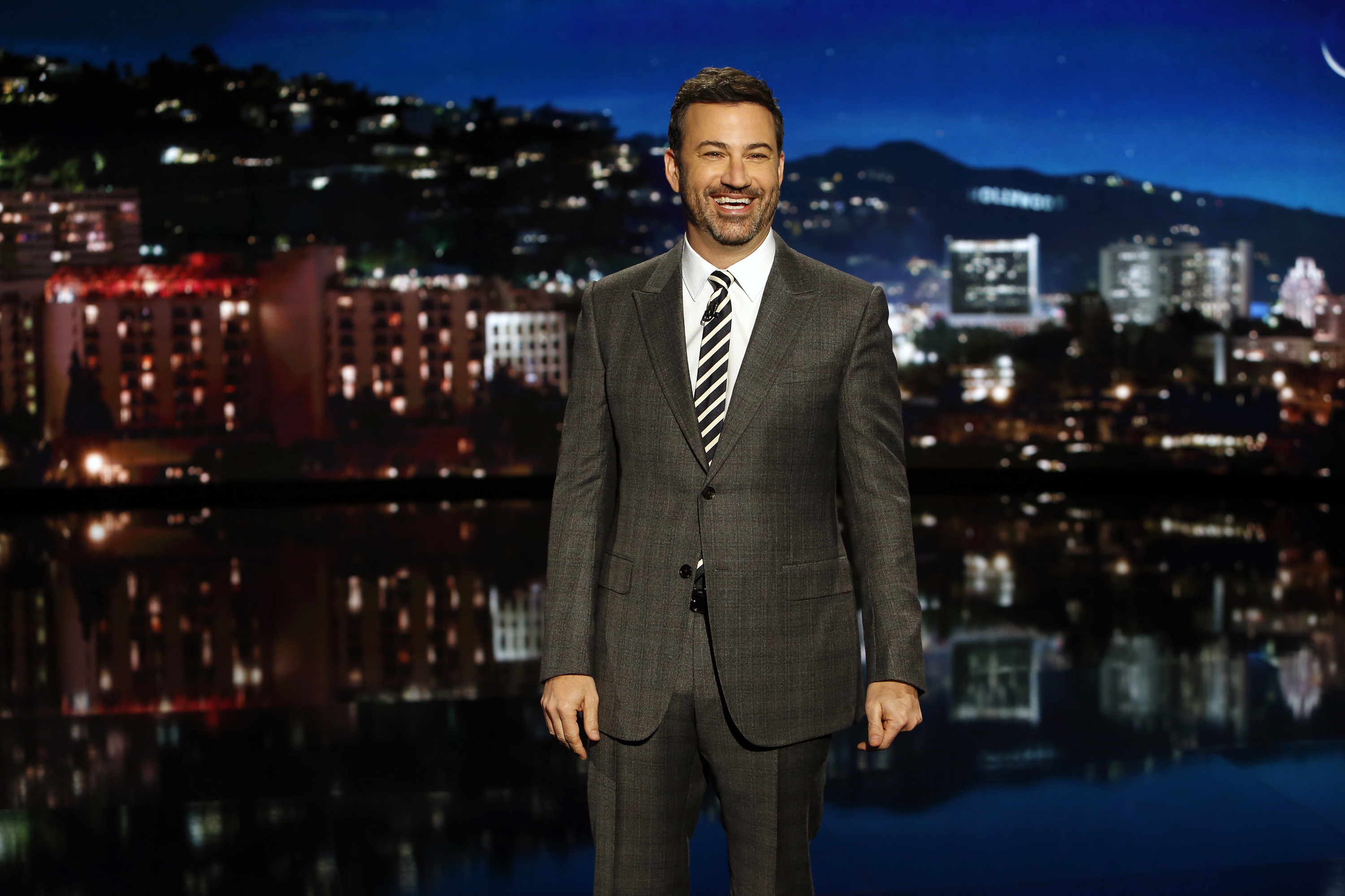 Jimmy Kimmel stands smiling in a gray suit against a backdrop of the Los Angeles skyline at night.