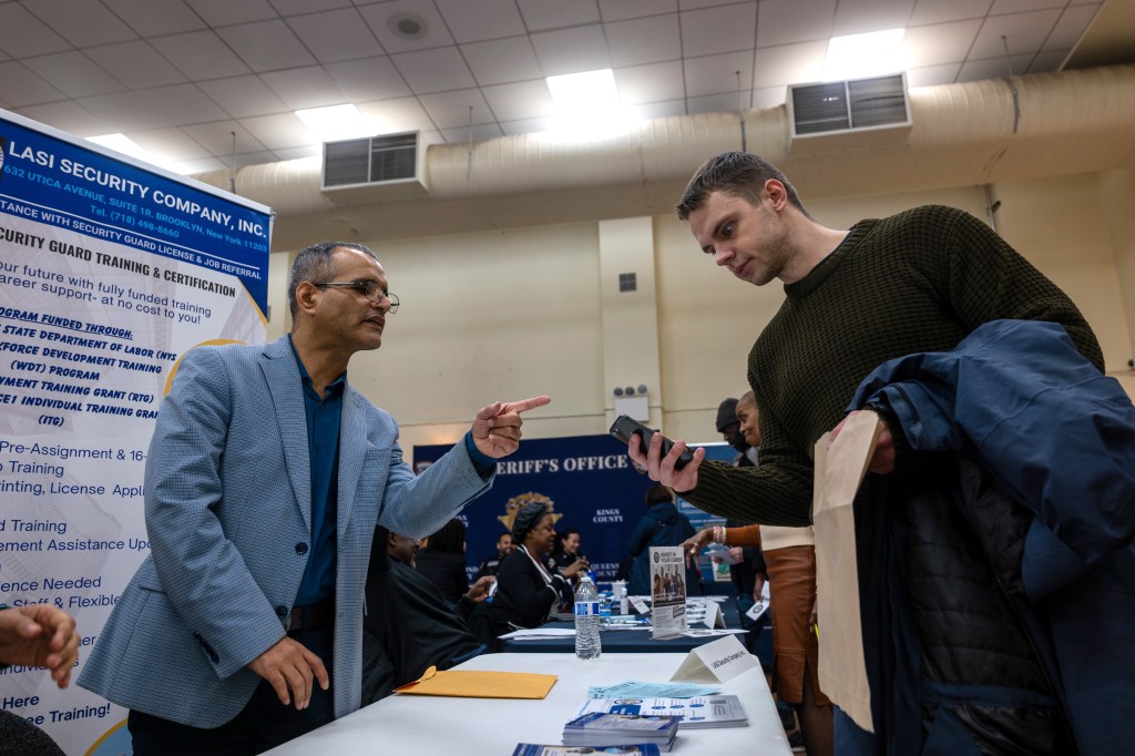 A man in a suit jacket gestures while speaking to a man holding a phone at a job fair.