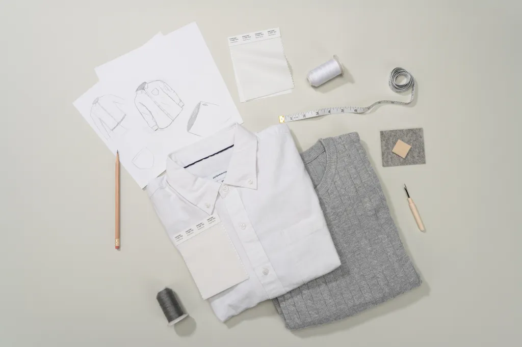 Fashion design flat lay with a white shirt, gray sweater, white and gray thread, fabric swatches for Pantone 11-4201 Cloud Dancer, sketches, measuring tape, and a seam ripper.