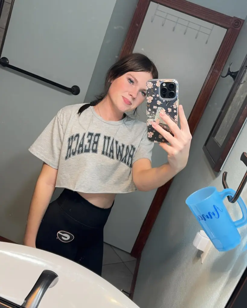 A young woman takes a mirror selfie in a cropped t-shirt and athletic pants.