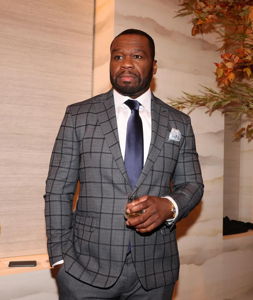 Curtis '50 Cent' Jackson wearing a grey windowpane plaid suit, white dress shirt, navy tie, and light blue pocket square, holding a glass of amber liquid.