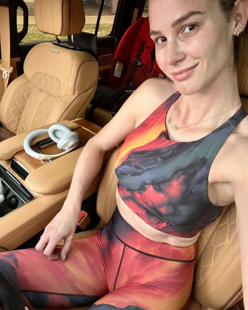Meghan King wearing a red, orange, and black tie-dye athletic top and matching leggings sits in the driver's seat of a car.