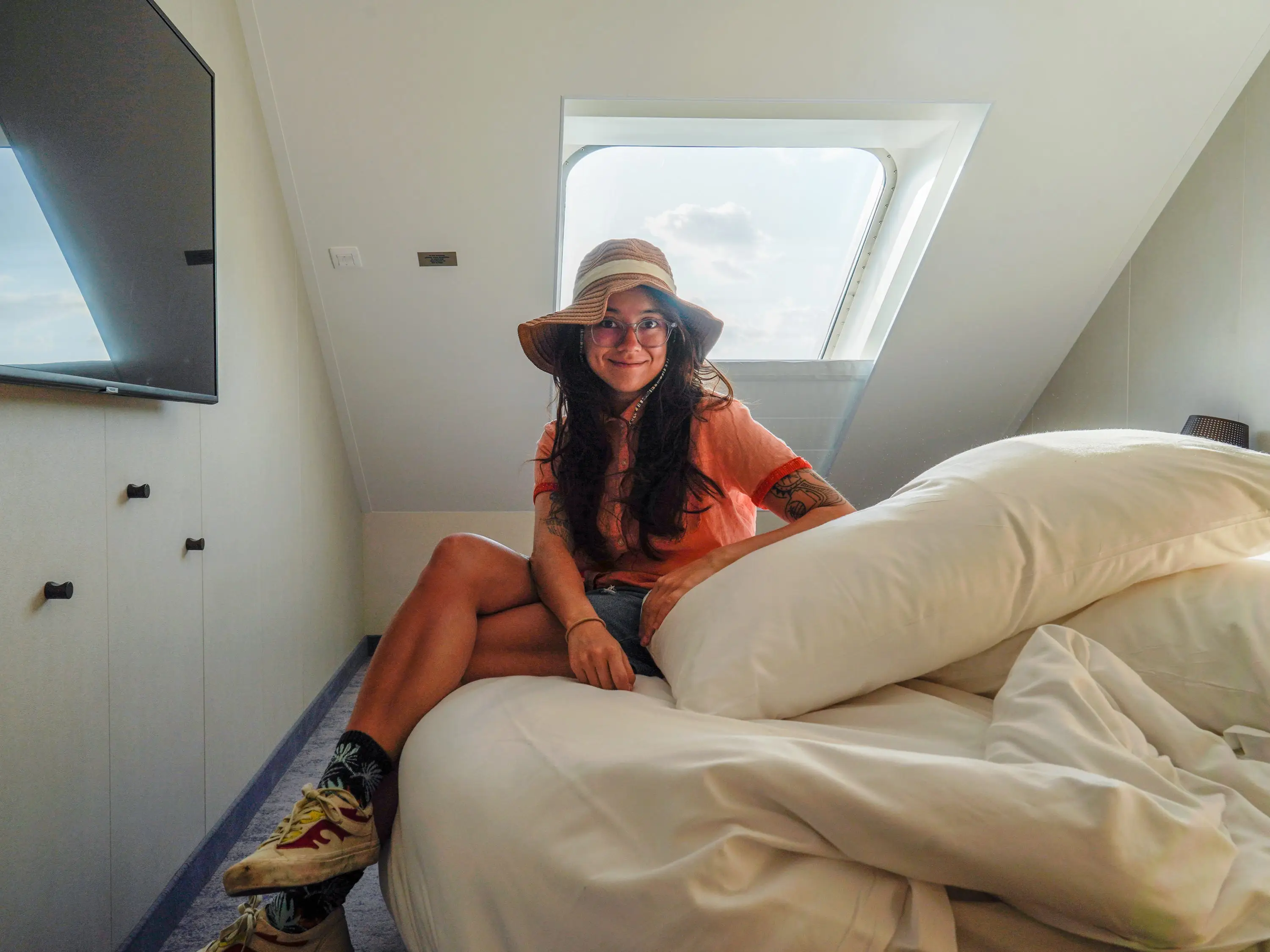The author, wearing a T-shirt, shorts, and a sun hat, sits on an unmade bed with white sheets in a stateroom.