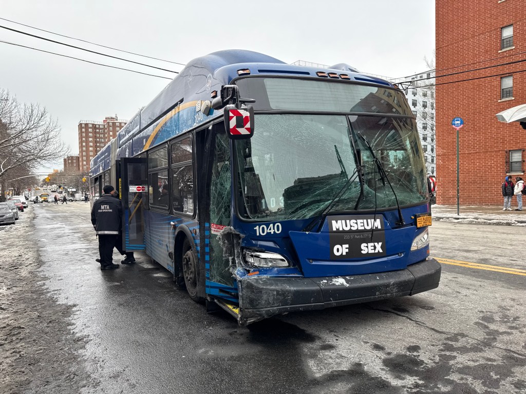 A damaged blue MTA bus with a shattered windshield and front bumper after a mechanical malfunction, with a person wearing an MTA Road Operations jacket standing by the open door, on a street with patches of snow and ice.