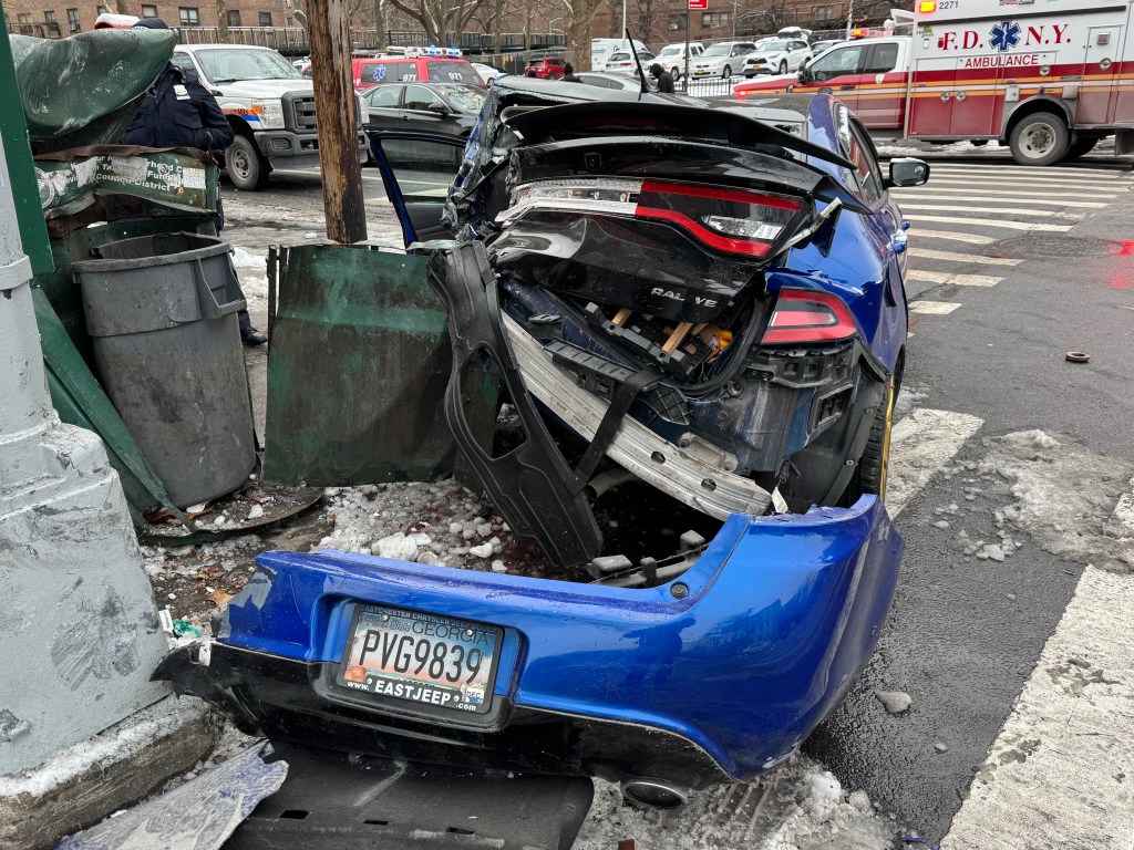A blue Dodge Charger, heavily damaged in the rear, rests next to crumpled green trash containers on a snowy street, with an FDNY ambulance visible in the background.