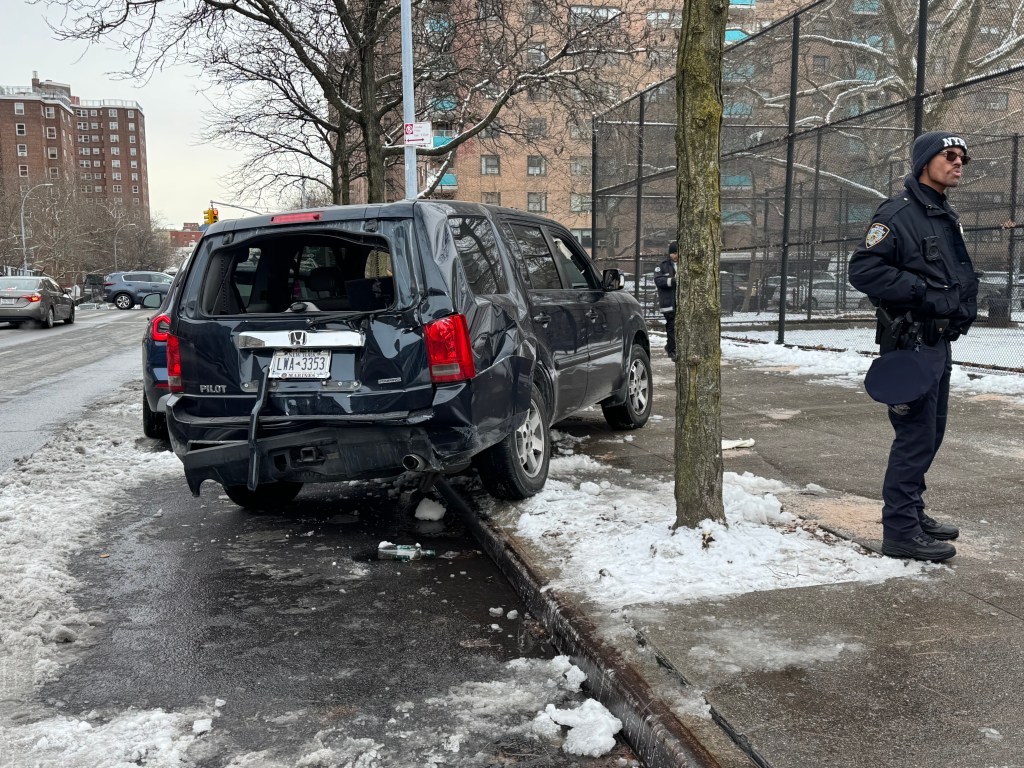 A black Honda Pilot, with severe damage to its rear and back window, is parked on the street with a police officer standing nearby.
