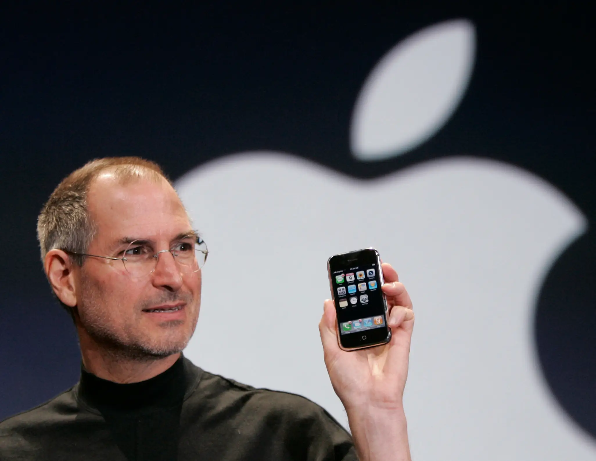 Former Apple CEO Steve Jobs shows off the first iPhone on stage at an Apple event with the Apple logo looming in the background.