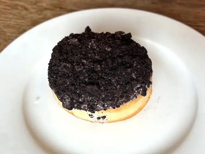 A doughnut with no hole topped with icing and crushed Oreos on a white plate.