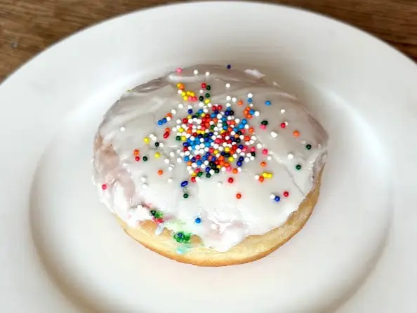 A doughnut with no hole topped with white icing and rainbow sprinkles on a white plate.