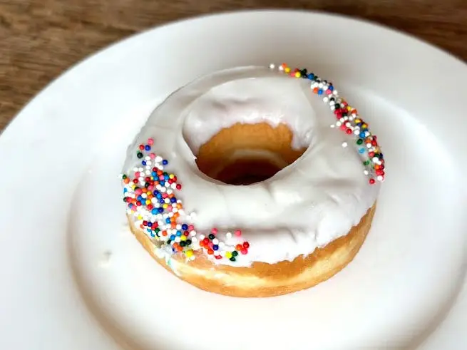 A doughnut topped with white icing and rainbow sprinkles on a white plate.