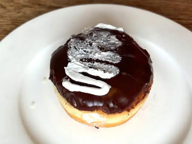 A doughnut with no hole topped with chocolate icing and a squiggle of white icing on a white plate.