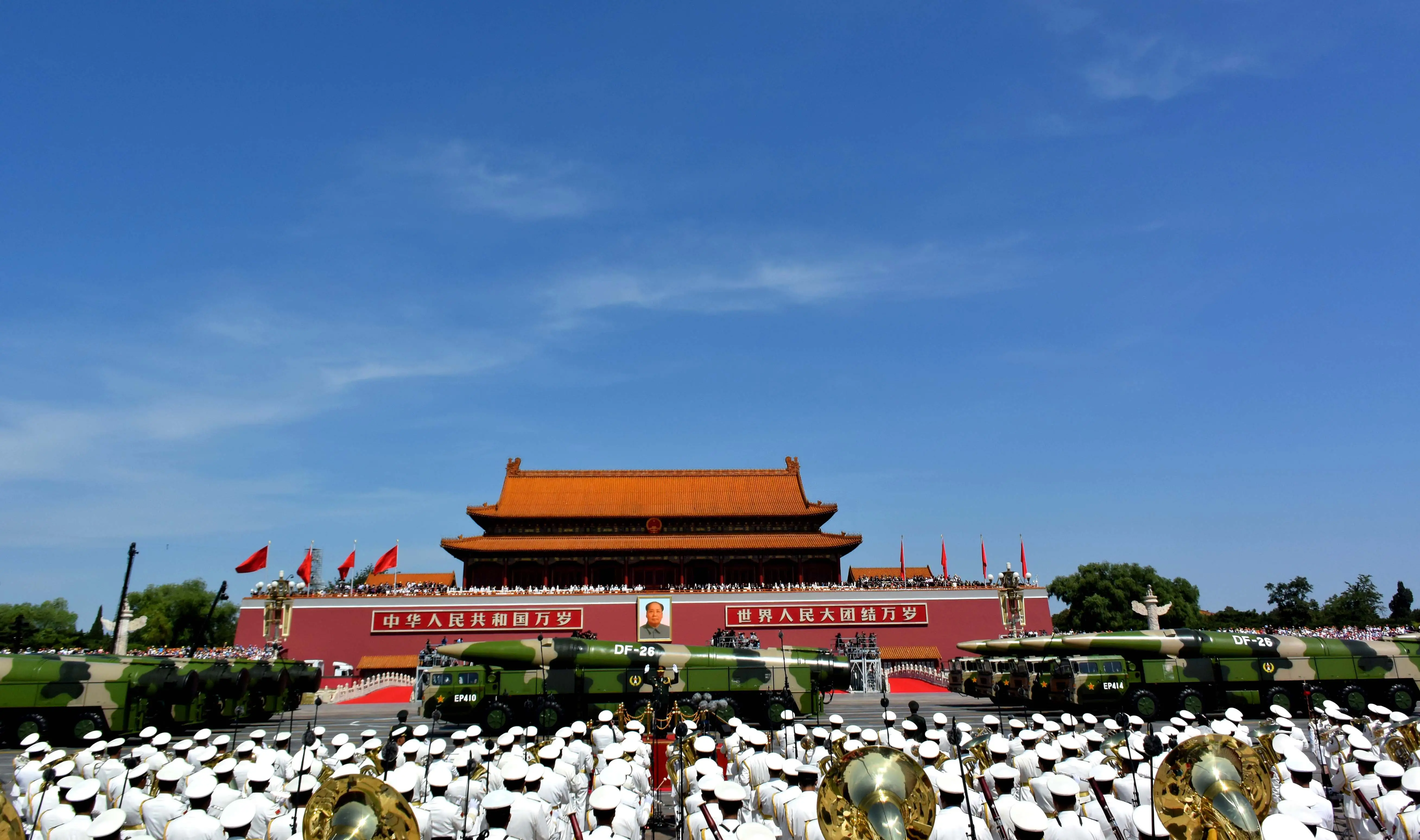 Chinese DF-26 missiles, camouflage colored, in front of military personnel standing at a parade against a blue sky.