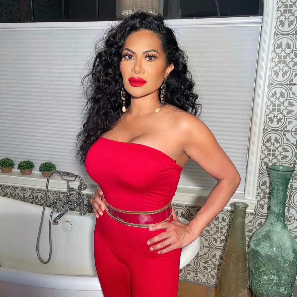 A woman in a red strapless jumpsuit and gold belt poses with her hands on her hips.