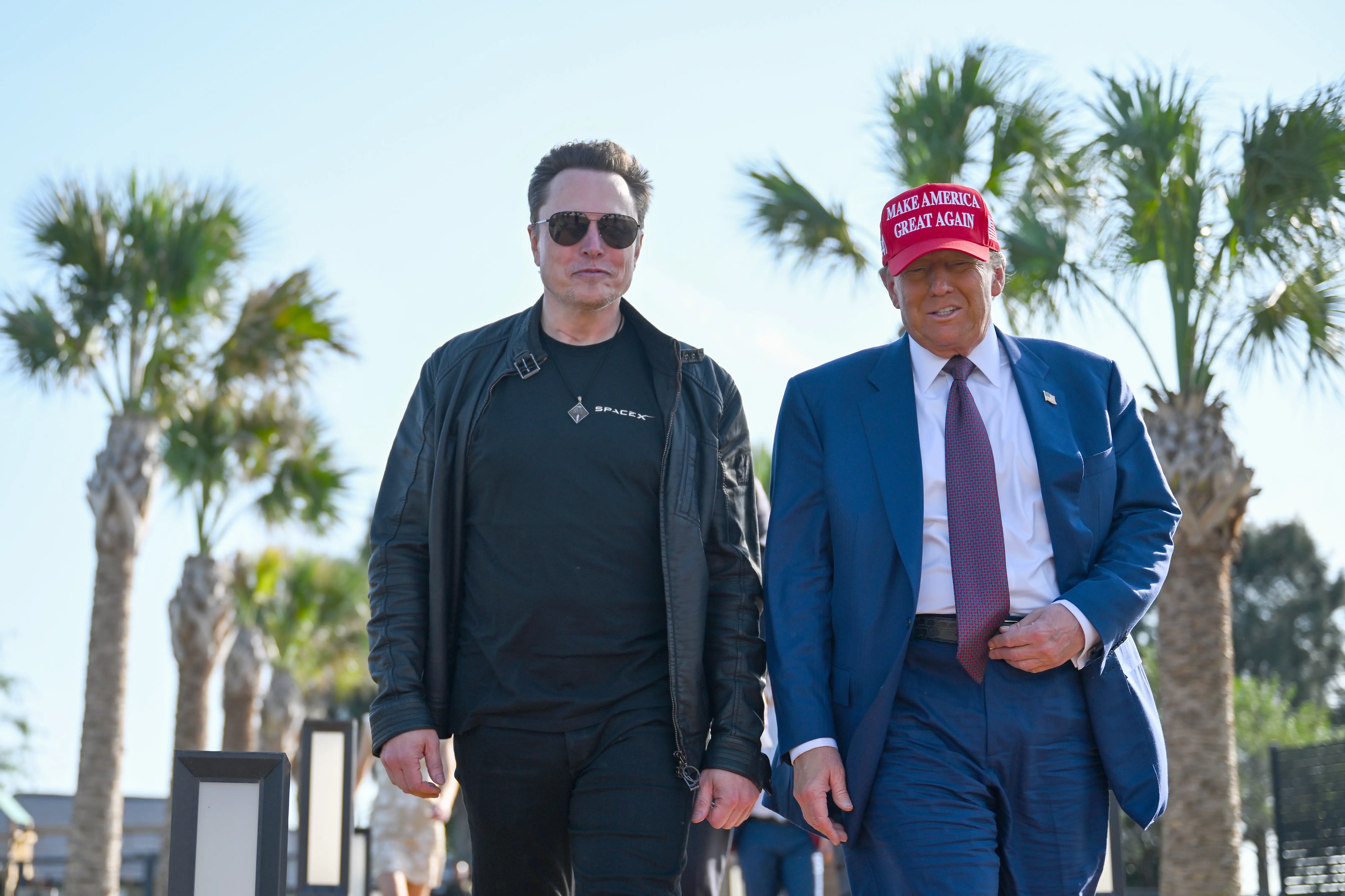 Elon Musk and Donald Trump walking together with palm trees behind them. Musk is wearing an all-black outfit with a SpaceX T-shirt, and Trump is wearing a suit and tie and a