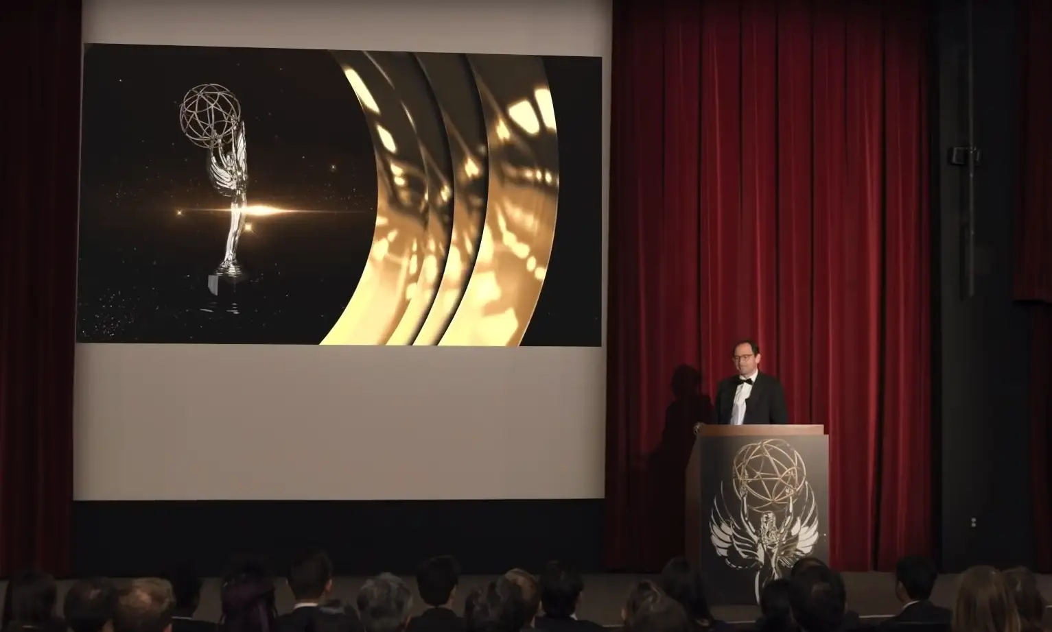 Jon Gray stands behind a podium at a fake award show, with a red carpet and screen behind him.