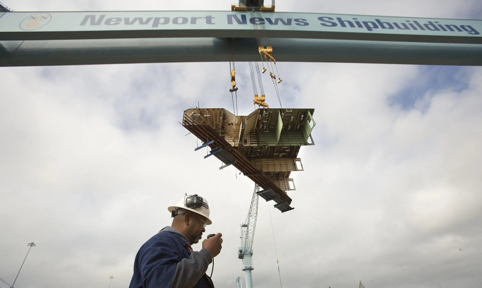 A man wearing a hard hat walks under a crane reading 