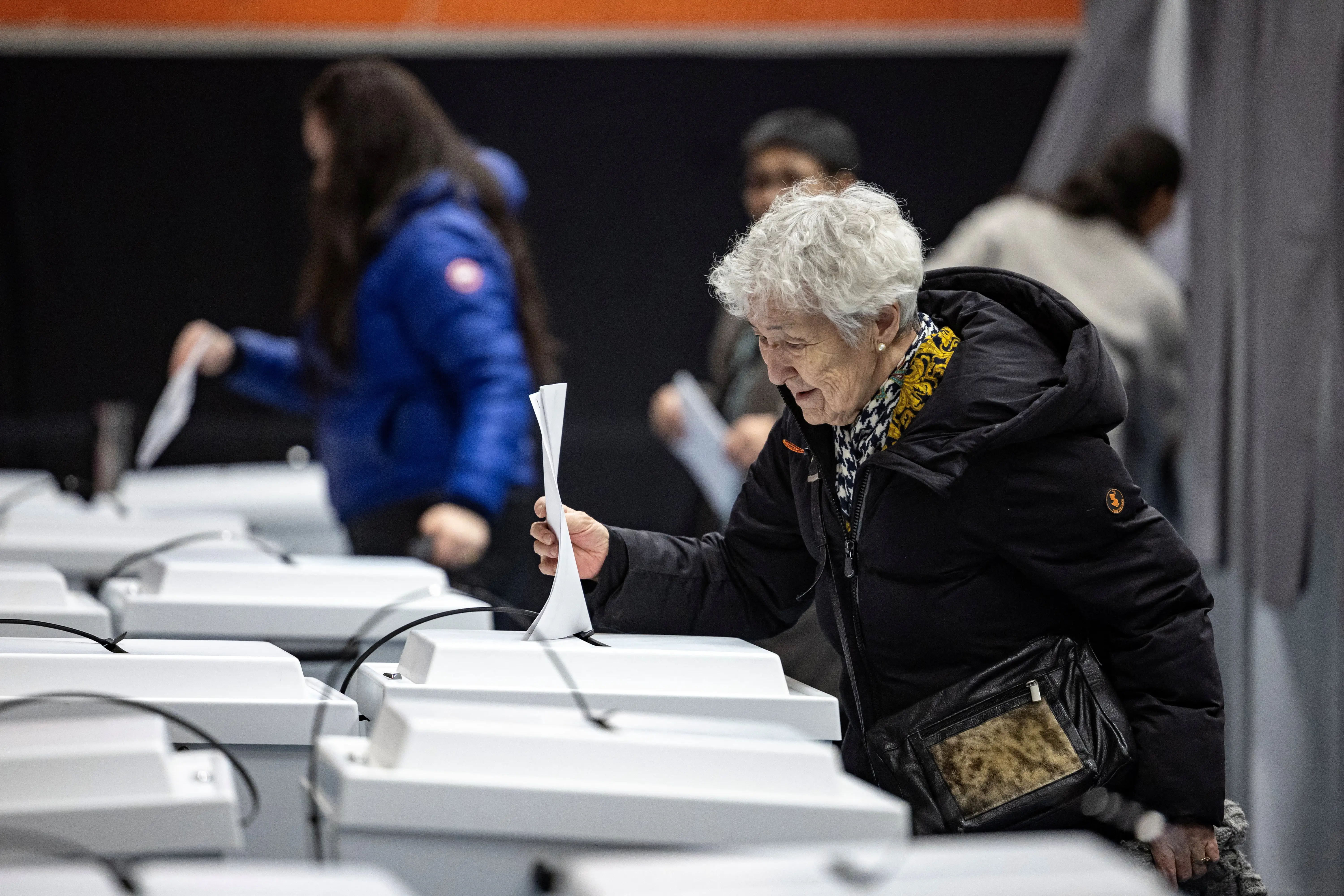 An elderly woman in a black winter coat casts a ballot dropping it into a large white box