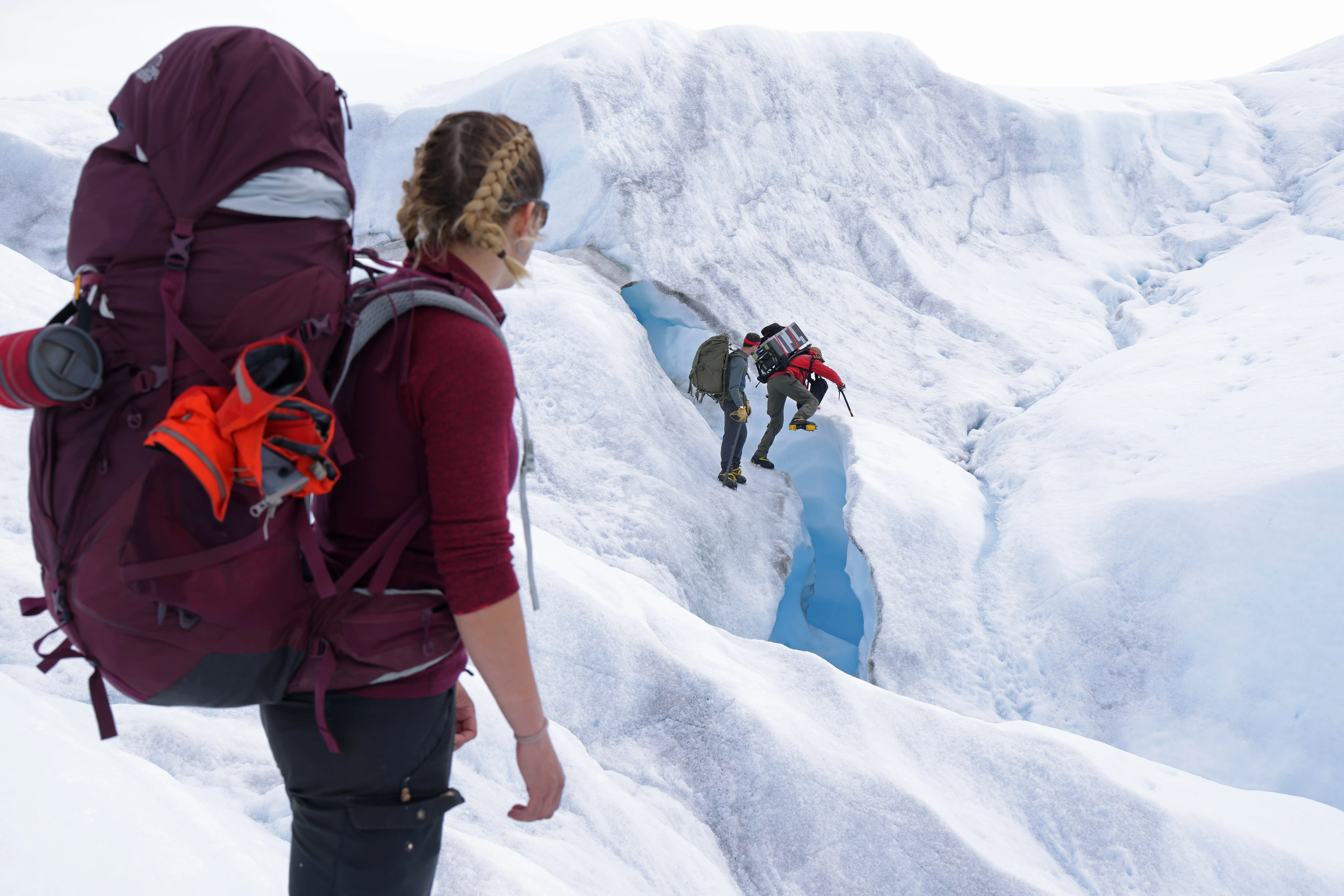 A woman with braids and a large backup looks at two people walking on a glacier