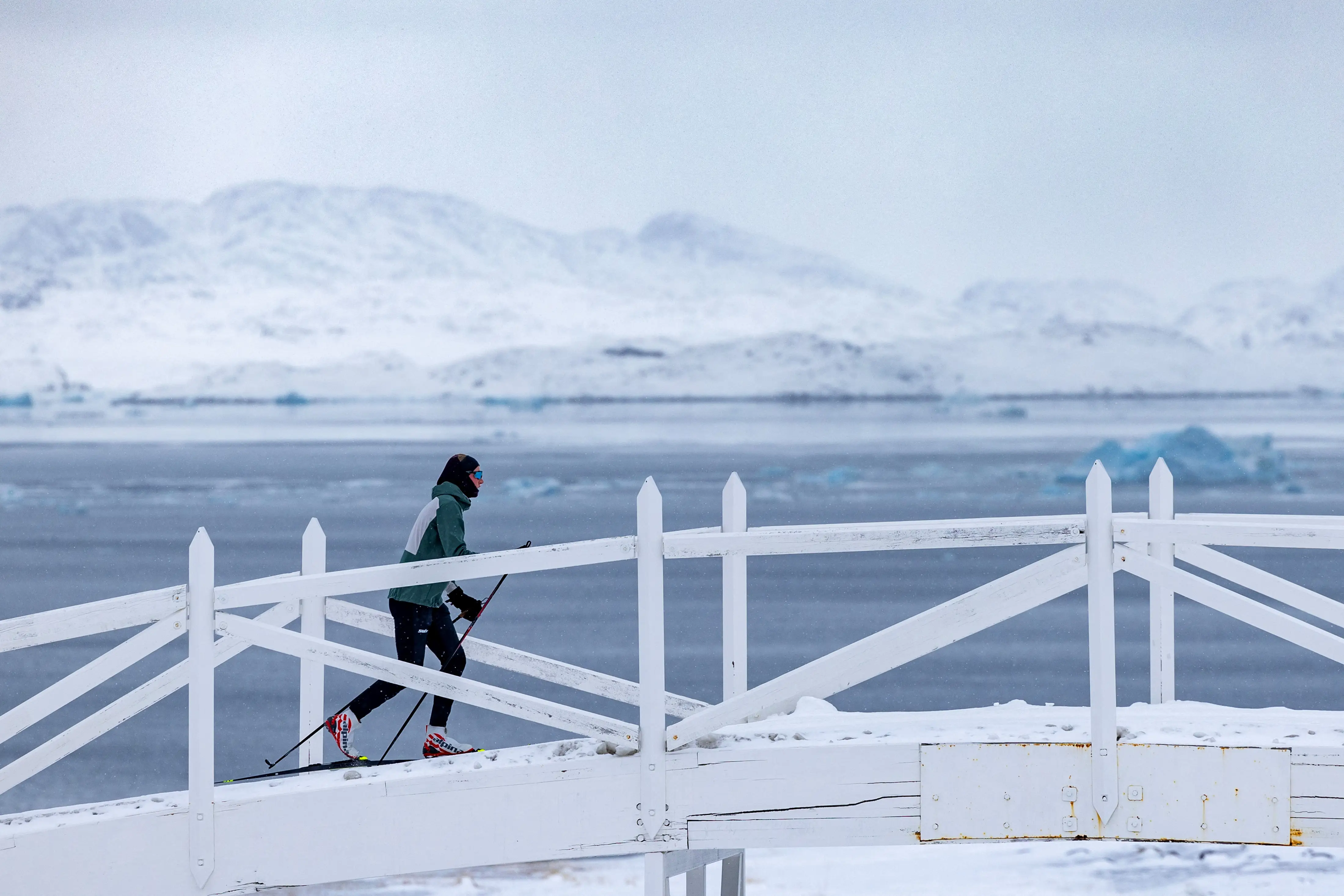 A cross country skier on a white bridge with snowy hills and water in the background